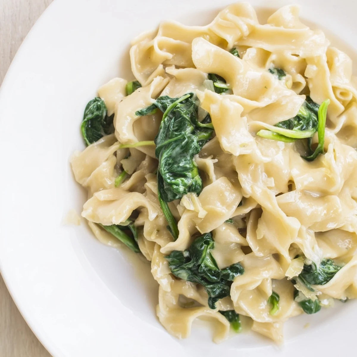 A close-up view of Creamy Spinach Garlic Noodles showcases glossy, garlicky cream sauce clinging to each wide noodle and vibrant green spinach.